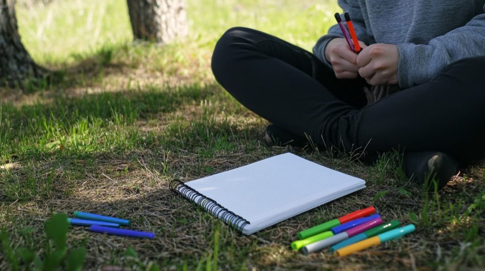 n outdoor setting where a person sits cross-legged on the grass with a blank notebook and a set of colorful markers nearby. ai generated